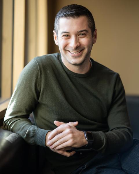 Head-and-shoulders portrait of a smiling person wearing a dark green sweater, seated indoors and facing the camera.