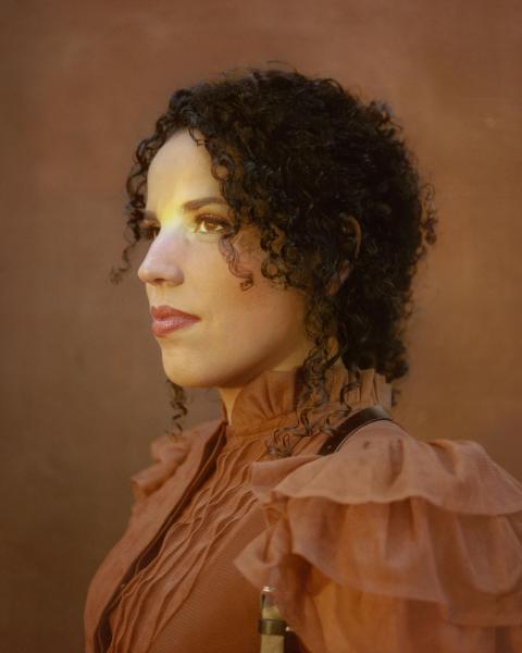 Profile view of a woman with curly dark hair wearing a ruffled blush-toned top, photographed against a warm brown background.