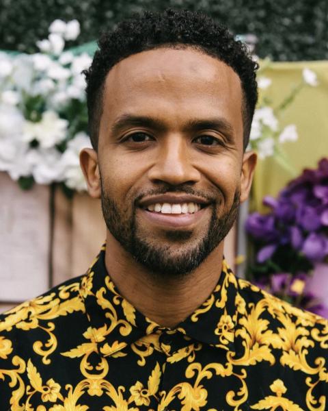 Headshot of a man smiling, wearing a black and gold patterned shirt, with greenery and flowers in the background.