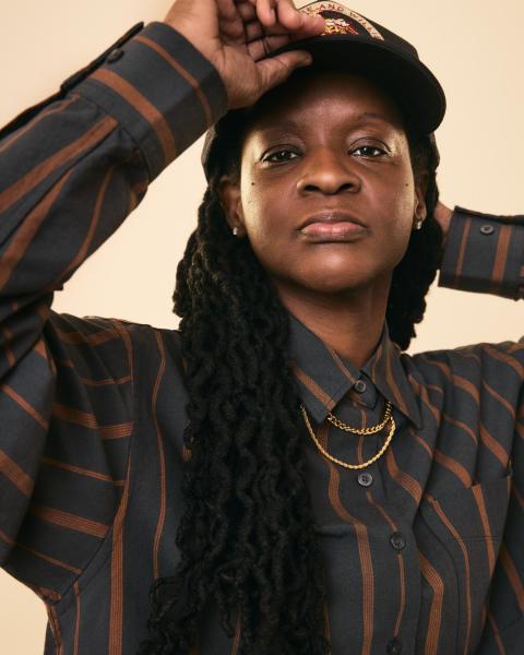 Person wearing a striped shirt and cap, long braided hair, gold necklaces, neutral background.