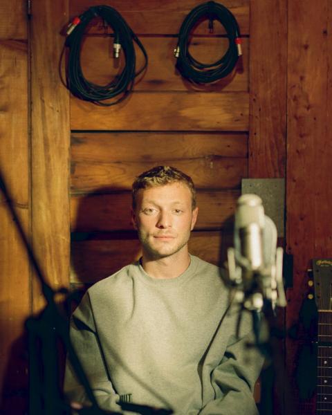 A young man with light hair sits in front of a wooden wall, wearing a grey sweatshirt. A studio microphone is positioned in the foreground, with a guitar and coiled audio cables visible in the background. 