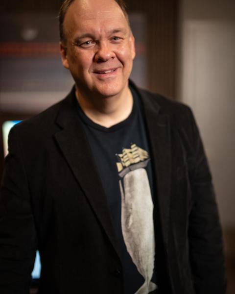 Man smiling, wearing a black blazer over a navy graphic t-shirt, photographed indoors against a softly lit background.