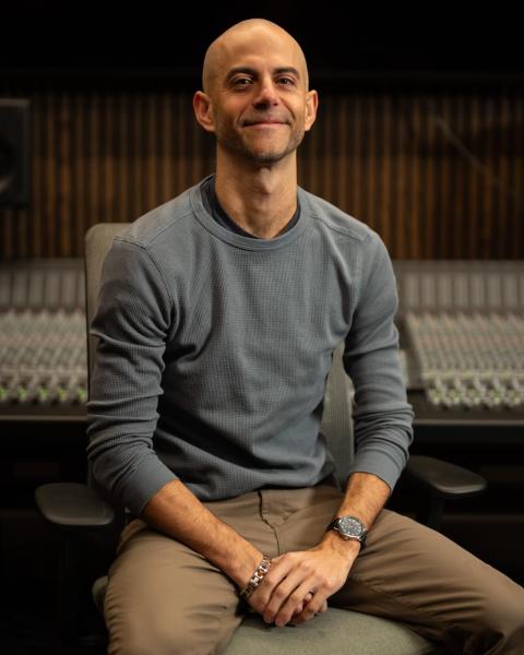 A bald man wearing a grey long-sleeve shirt sits with his hands folded, smiling at the camera. Behind him is a large mixing console and studio monitor speakers in a professional recording studio setting.