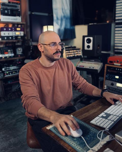Man sitting at a studio desk working at a keyboard and mouse, surrounded by professional audio equipment and studio monitors.