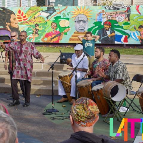 A vibrant mural serves as the backdrop while a group of individuals enthusiastically play drums and sing together, creating a harmonious and lively atmosphere.