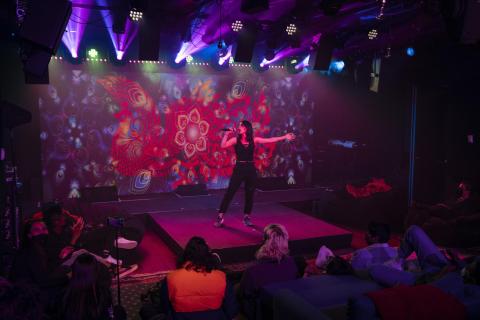 A performer poses and sings onstage in front of an audience, backed by an LED wall with elaborate visuals, as lights and projections are created.