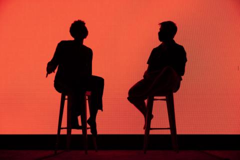 Two performers are sitting, silhouetted in front of an LED wall glowing orange.