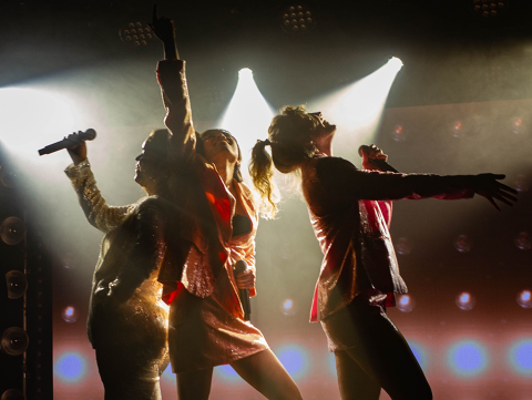 Three singers pose with mics illuminated by stage lighting in the Black Box Theater at BerkleeNYC.