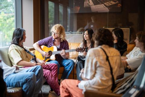 A group of students sit together in a casual lounge setting while a person with curly blonde hair plays acoustic guitar. The group appears engaged in conversation and collaboration, with music equipment and a vintage amplifier visible in the background.