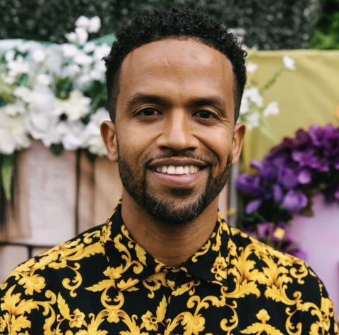 Headshot of a man smiling, wearing a black and gold patterned shirt, with greenery and flowers in the background.
