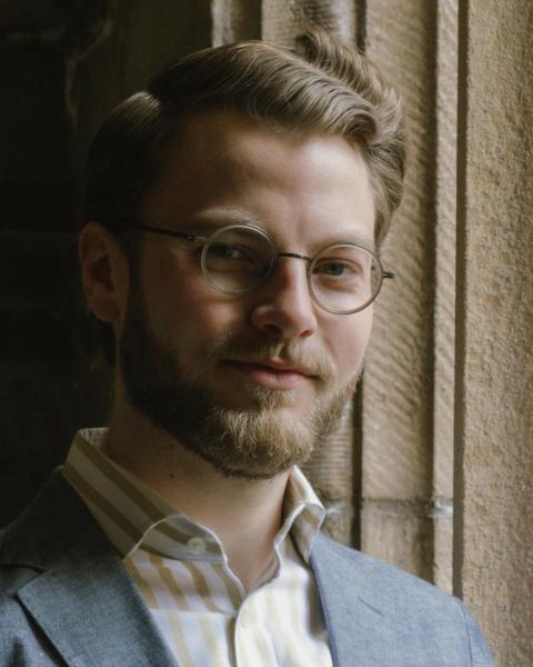 Man wearing round wire-framed glasses and a light blue blazer over a striped shirt, photographed against a stone wall backdrop