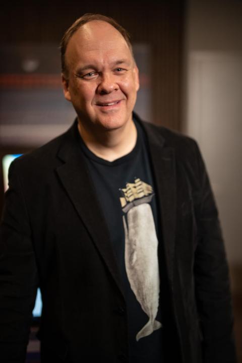Man smiling, wearing a black blazer over a navy graphic t-shirt, photographed indoors against a softly lit background.