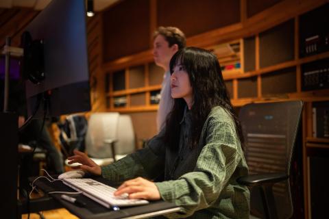 A young woman with long dark hair sits at a computer workstation in a dimly lit studio environment, focused on the screen. A second person is visible in the background.