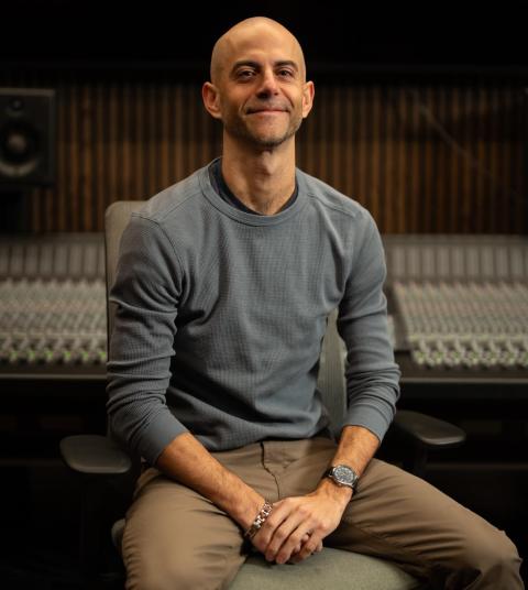 A bald man wearing a grey long-sleeve shirt sits with his hands folded, smiling at the camera. Behind him is a large mixing console and studio monitor speakers in a professional recording studio setting.