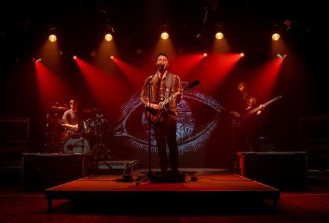 A guitarist and vocalist performs at center stage under dramatic red lighting, flanked by a drummer and bassist. A large eye graphic is projected onto the backdrop behind them.