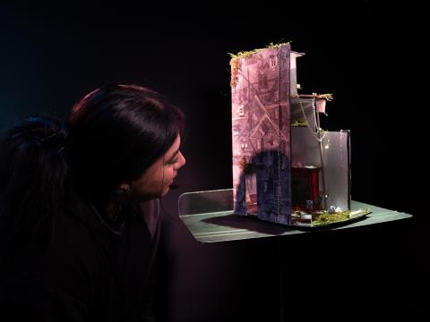 A person with dark hair leans in closely to examine a detailed miniature architectural model, illuminated against a dark background. The intricate model features small buildings, moss, chains, and tiny decorative elements.